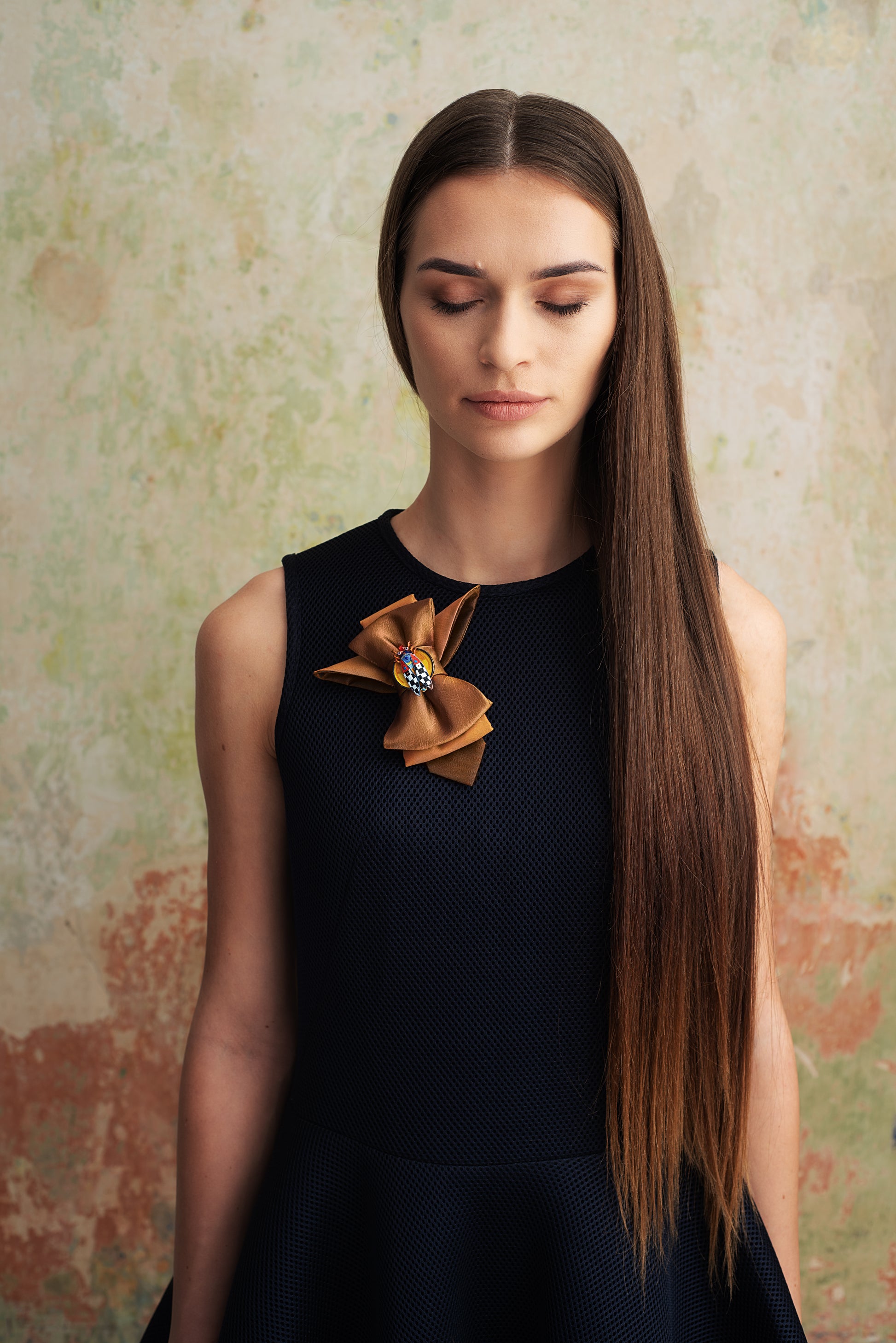 Woman wearing a black dress with a decorative brooch against a textured wall handmade luxury neck accessory, women statement tie, vertical bow tie for women, gold satin necktie, elegant women tie, unique bow tie alternative, luxury handmade tie, artistic neckwear for women, modern formal accessory, gold statement necklace tie, designer women necktie, wedding guest accessory women, avant garde fashion tie, silk bow tie alternative, bold women fashion accessory, golden necktie women, formal outfit statement p