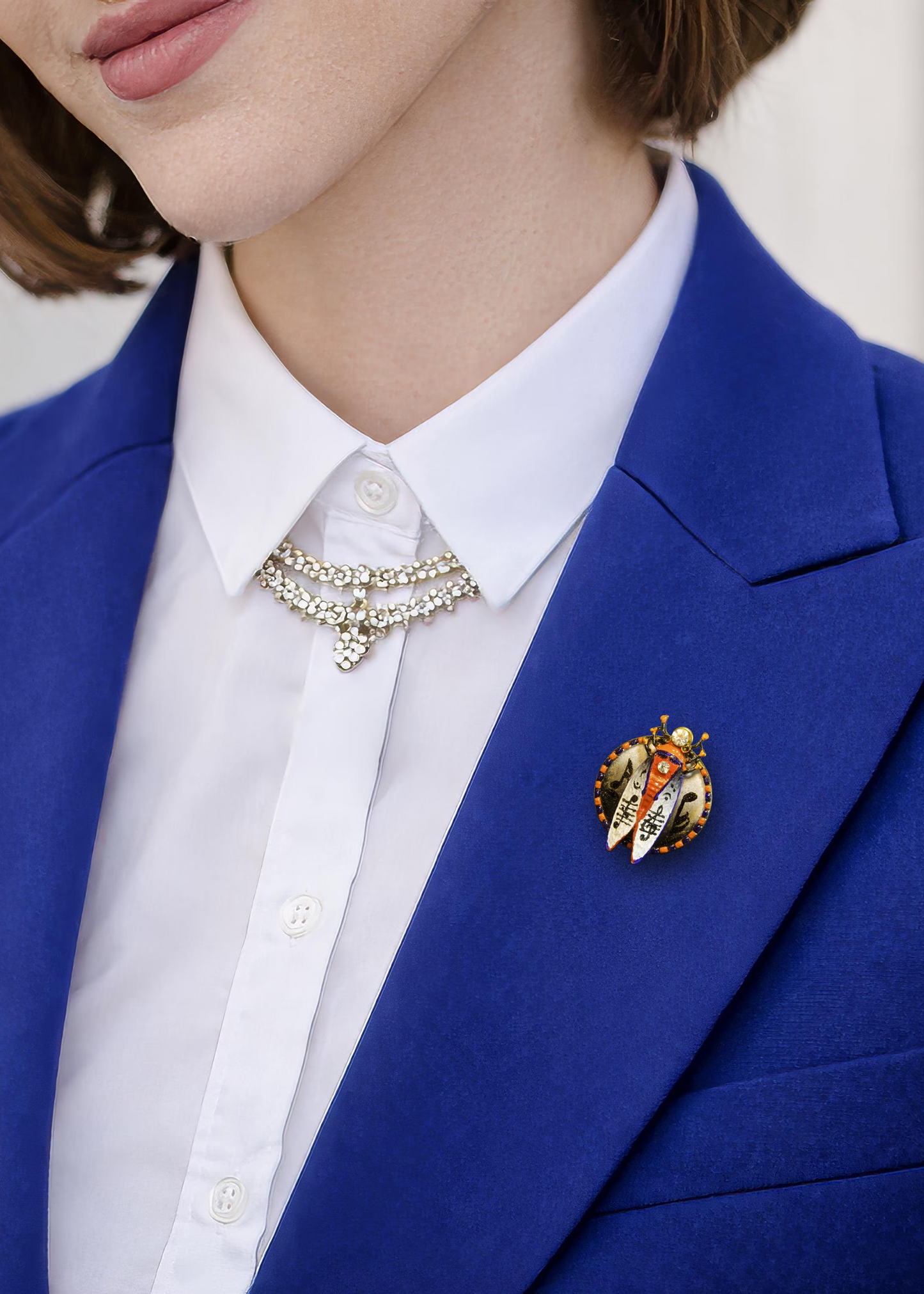 Person wearing a blue blazer with a white shirt and decorative brooch hand painted insect brooch, enamel beetle brooch, cicada brooch jewelry, rhinestone insect brooch, colorful bug brooch, artistic insect pin, handmade enamel brooch, statement insect jewelry, unique bug brooch, designer insect brooch, red black white beetle brooch, decorative insect pin, nature inspired brooch, collectible insect jewelry, whimsical bug brooch, vintage style insect brooch, artistic enamel insect pin, crystal insect brooch, 