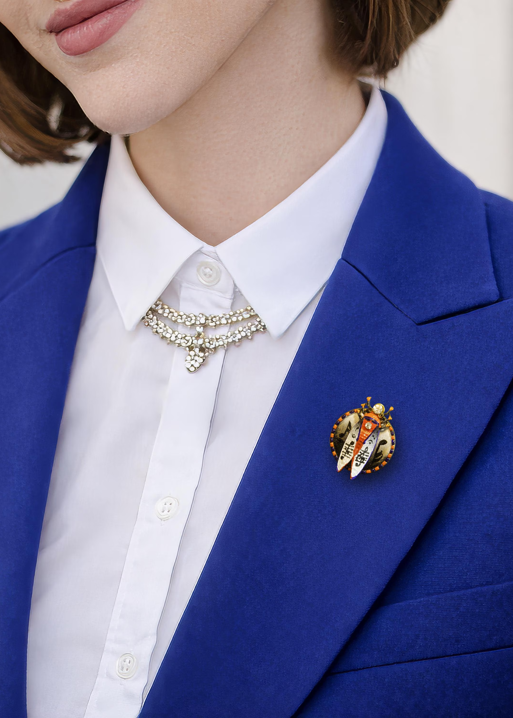Person wearing a blue blazer with a white shirt and decorative brooch hand painted insect brooch, enamel beetle brooch, cicada brooch jewelry, rhinestone insect brooch, colorful bug brooch, artistic insect pin, handmade enamel brooch, statement insect jewelry, unique bug brooch, designer insect brooch, red black white beetle brooch, decorative insect pin, nature inspired brooch, collectible insect jewelry, whimsical bug brooch, vintage style insect brooch, artistic enamel insect pin, crystal insect brooch, 