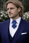 Man wearing a blue suit with a patterned tie against a blurred floral  Man wearing a navy suit with a white shirt and blue patterned tie, standing against a blurred floral background blue champagne hopper tie, artistic vertical bow tie for men, unique groom accessory wedding tie, elegant blue textured statement neckwear, modern alternative bow tie, handmade designer tie Lithuania, stylish groom wedding accessory, avant garde men’s formal neckwear, luxury artistic tie for special occasions, blue chbackground