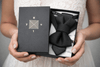 Person holding a box with black bow ties and 'RYU' branding Handmade black hopper tie for wedding, original designer bow tie on white shirt, Luxury black wedding hopper tie, modern bow tie and necktie hybrid, Elegant handmade hopper tie for groom, minimalist black formal accessory, Original design black hopper tie, premium wedding bow tie for men, Modern black hopper tie for formal wear, handcrafted wedding neck accessory, Designer handmade hopper tie, sophisticated black tie