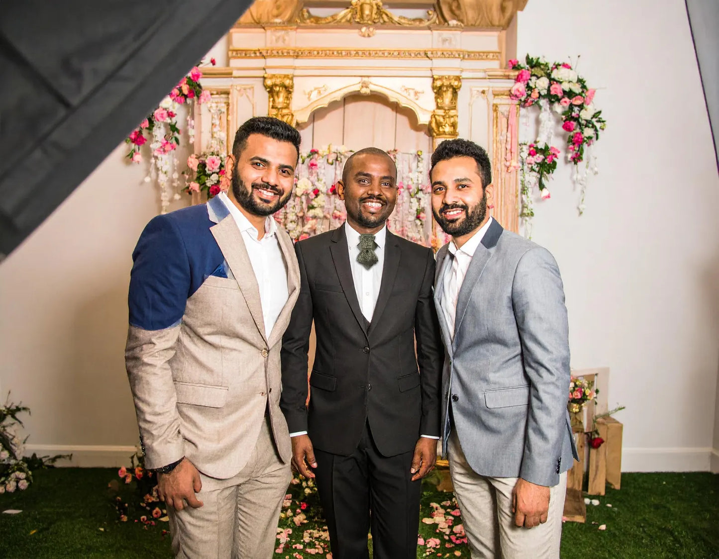 Three men posing together in front of a decorated wall with floral A black and gold ascot tie with a detailed paisley pattern, worn with a classic white dress shirt. The tie features a chevron stripe texture, creating a sophisticated and formal look. It's a handmade luxury wedding accessory arrangements.