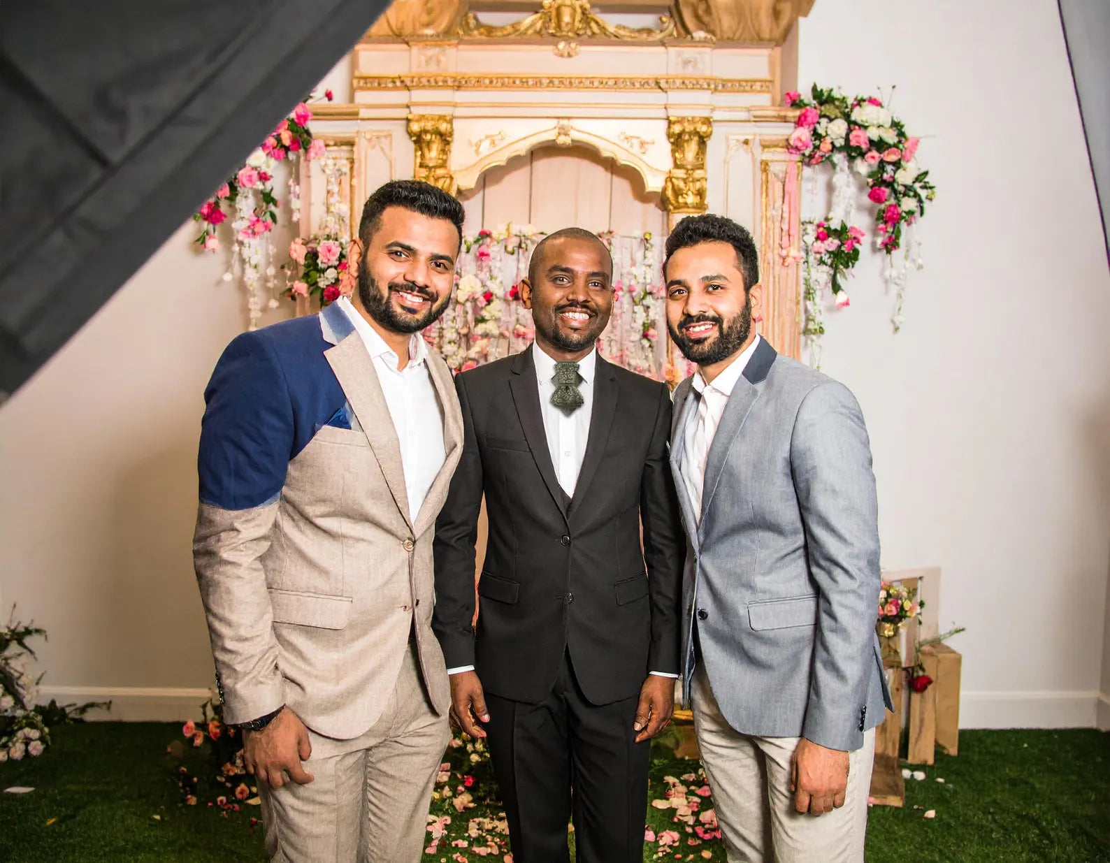 Three men posing together in front of a decorated wall with floral A black and gold ascot tie with a detailed paisley pattern, worn with a classic white dress shirt. The tie features a chevron stripe texture, creating a sophisticated and formal look. It's a handmade luxury wedding accessory arrangements.