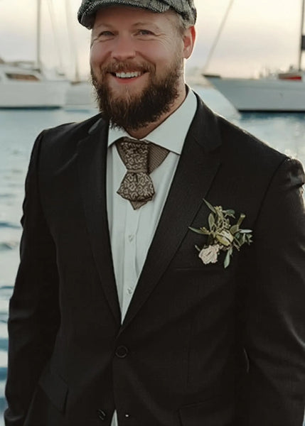 Man in formal attire with a boutonniere standing by water Bow Tie, Tie for wedding suite SAFARI hopper tie Bow tie 
A black and gold silk ascot tie with a detailed paisley pattern, worn with a classic white dress shirt. The tie features a chevron stripe texture, creating a sophisticated and formal look. It's a handmade luxury wedding accessory