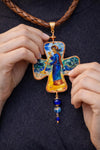 Cross-shaped pendant with artistic design held by hands against a dark Decorative cross-shaped pendant with a figurine on a dark background Unique artistic pendant for elegant woman, Hand painted miniature art pendant for women one of a kind jewelry, Luxury OOAK miniature painting necklace close up, Original artwork pendant by artist Marijus Piekuras, Fine art wearable pendant handcrafted detail, Tiny original painting inside pendant frame, Woman wearing one of a kind miniature pendant