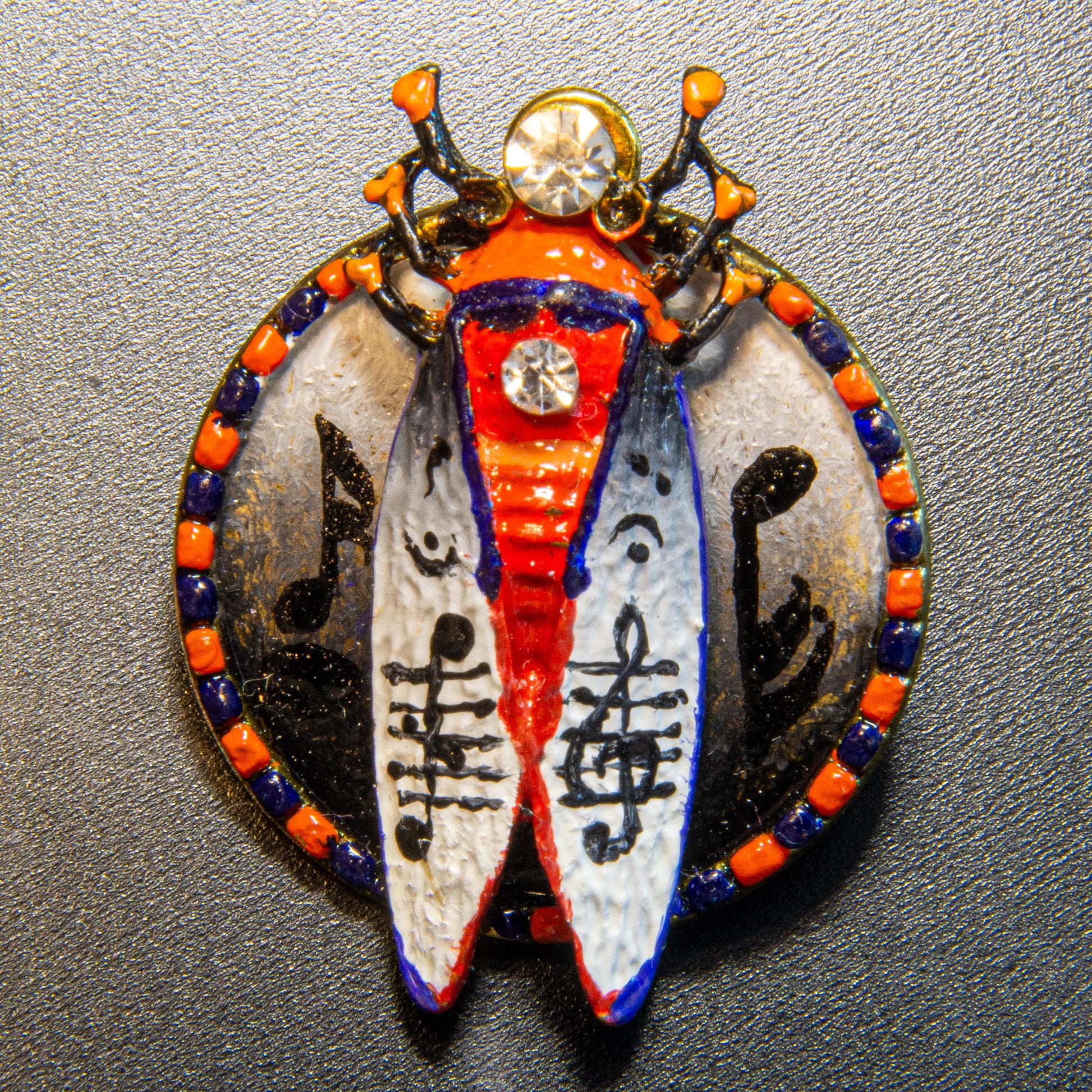Decorative brooch with a colorful design on a gray background Musical scarab brooch with hand painted music notes, unique artist beetle pin white background, handmade luxury music themed brooch, original art jewelry for musician gift, one of a kind painted insect brooch, red blue gold statement accessory, wearable art scarab with notes motif, artistic music lover brooch gift, handcrafted collectible beetle pin, exclusive small batch art jewelry