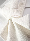 A close-up of a textured cream-colored necktie tied in a bow, displayed on a white collared shirt Luxury handmade white women's neck accessory featuring textured couture fabric, a satin ribbon bow, gold choker, and elegant pearl pendant. Close-up of a premium artisan statement necklace displayed on a jewelry bust, perfect for brides, weddings, bridal fashion, formal occasions, and timeless designer style. Sophisticated handcrafted textile jewelry with modern elegance and couture-inspired details.