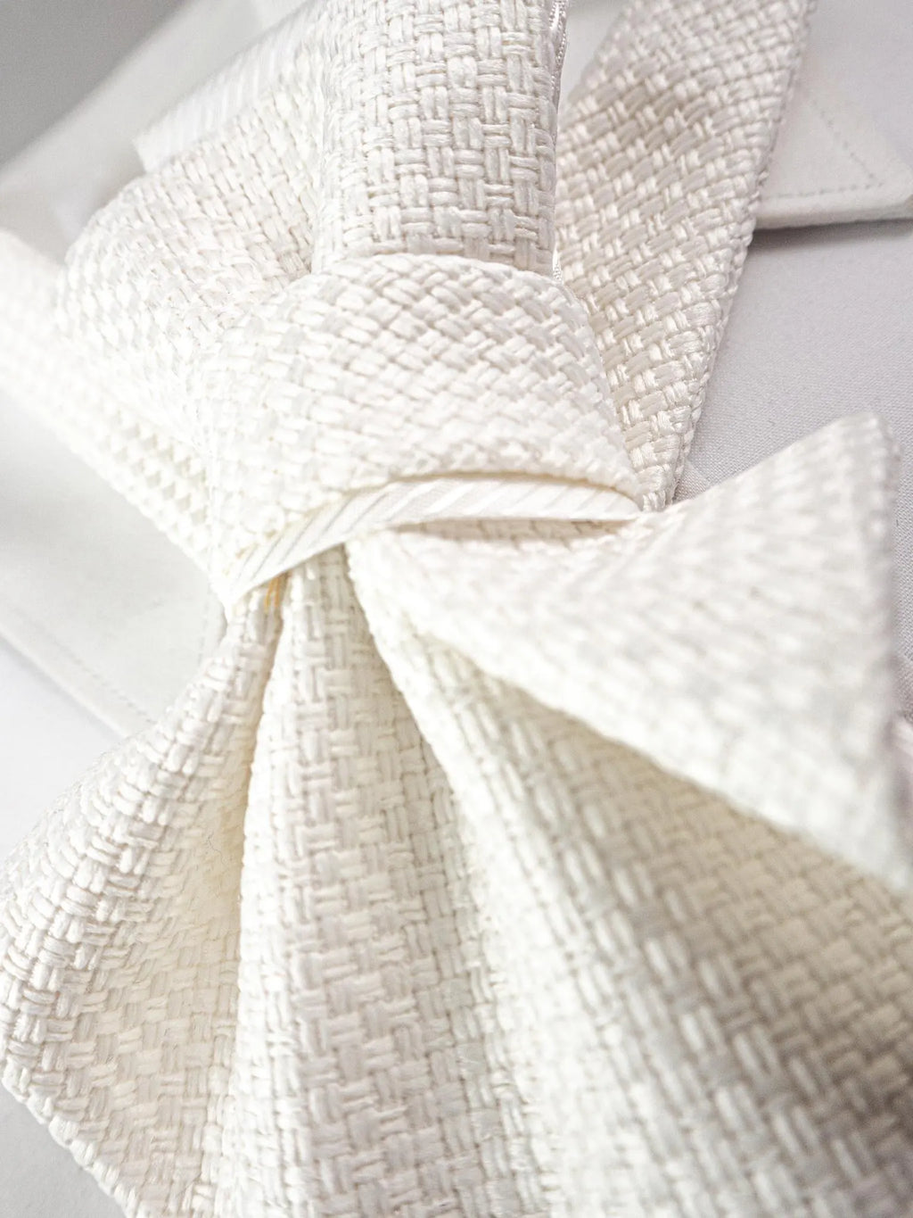 A close-up of a textured cream-colored necktie tied in a bow, displayed on a white collared shirt Luxury handmade white women's neck accessory featuring textured couture fabric, a satin ribbon bow, gold choker, and elegant pearl pendant. Close-up of a premium artisan statement necklace displayed on a jewelry bust, perfect for brides, weddings, bridal fashion, formal occasions, and timeless designer style. Sophisticated handcrafted textile jewelry with modern elegance and couture-inspired details.