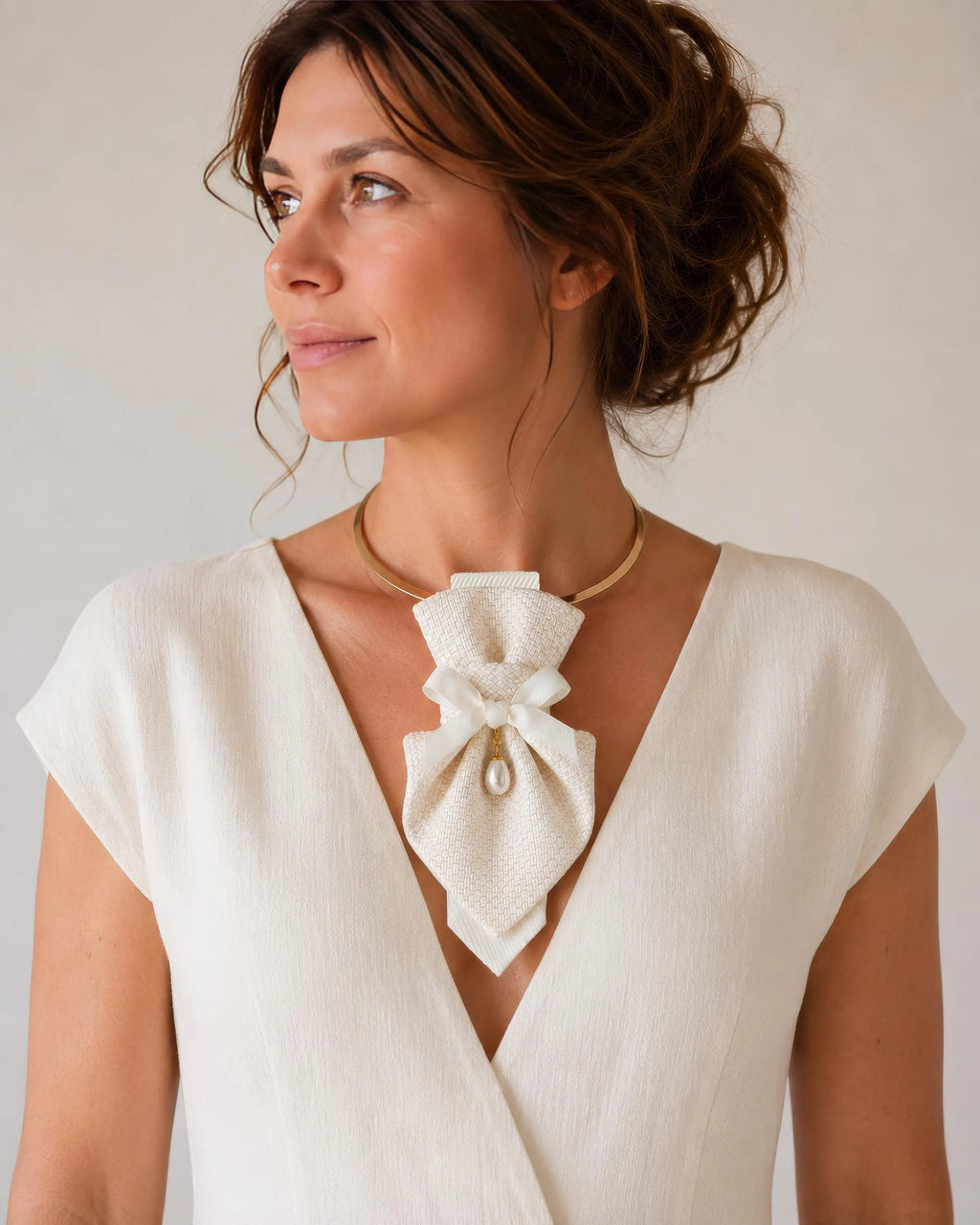 A woman wearing a cream-colored textured bow tie-neck accessory with a small pearl accent, styled over a white wrap dress against a neutral beige background Luxury handmade women's neck accessory featuring an elegant white textured fabric design on a gold choker with a satin bow and pearl pendant. Sophisticated bridal statement necklace for weddings, formal events, and special occasions. Artisan crafted fashion accessory with timeless elegance, perfect for brides, modern women, and unique designer style lov