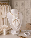A cream textured fabric bow with a pearl accent, displayed on a white mannequin bust. The bust is set on a wooden table surrounded by sewing tools, with a rack of lace dresses and fashion sketches in the background.