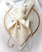 A gold-toned circular collar bar with a cream woven fabric bow, ribbon, and small pearl pendant, displayed on a white shirt collar White Hopper Tie with pearl pendant, handmade bridal neck accessory, textured white fabric tie, elegant ribbon bow, luxury wedding neckwear for women, white collared shirt, romantic bridal fashion, unique designer tie, pre-tied formal accessory, pearl necktie, wedding gift for bride, luxury handmade fashion accessory
