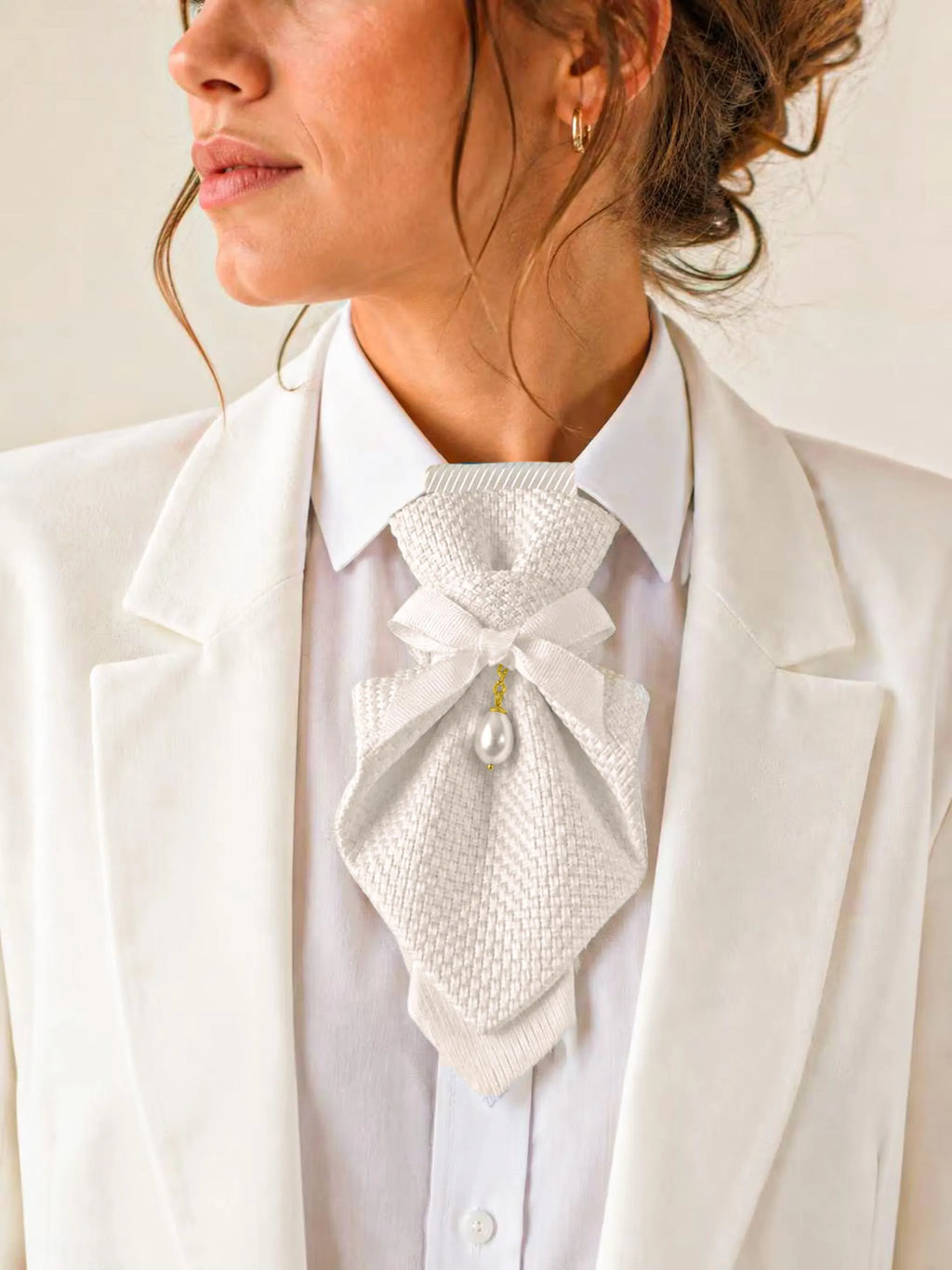 A close-up of a woman wearing a white textured bow tie with a single pearl pendant, styled over a white collared shirt and white blazer A white textured ascot tie featuring a central white ribbon bow and a single dangling white pearl pendant. The ascot is worn over a crisp white collared shirt and underneath a white blazer, creating a layered and formal appearance.