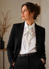 A woman wearing a black tailored blazer and trousers with a white dress shirt and a large white bow tie, posed indoors against a neutral beige wall white ascot tie, ivory wedding neckwear, elegant woman in black suit, white button-down shirt, pearl brooch accessory, bridal fashion accessory, handmade wedding tie, formal feminine neckwear