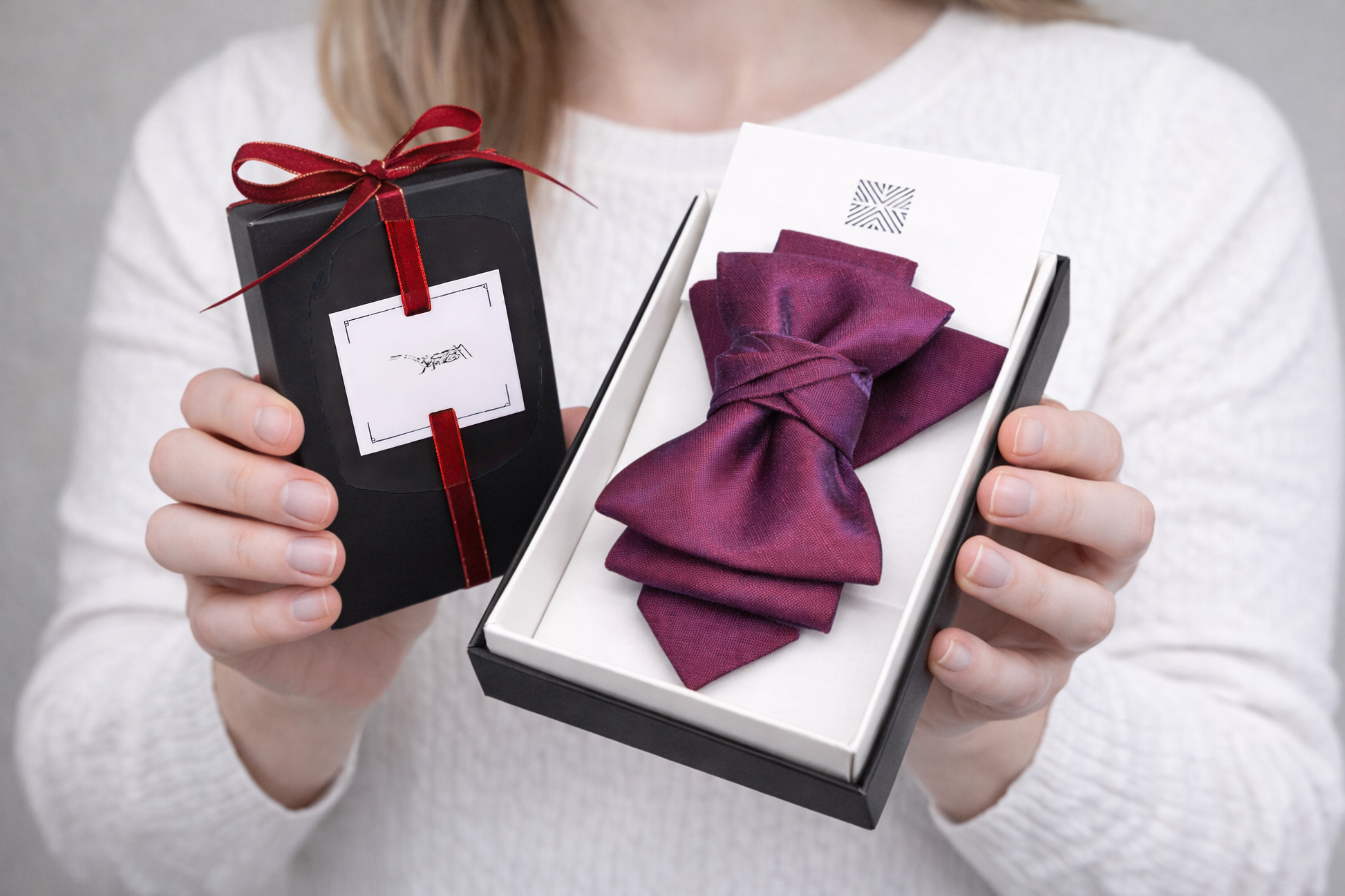 Person holding a black gift box with a red ribbon and a purple bow tie in an open box.