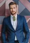 Man in a blue suit with a green bow tie standing against a geometric patterned wall  Luxury handmade jabot tie in green patterned fabric, decorated with Gediminas Columns and tricolor detail, unisex accessory for weddings, concerts, formal occasions, Heritage-inspired handmade tie in jabot style, green design with golden Gediminas Pillars and Lithuanian tricolor ribbon, symbol of identity and style, Elegant handmade necktie in green with Gediminas Columns emblem and tricolor detail, perfect for cult